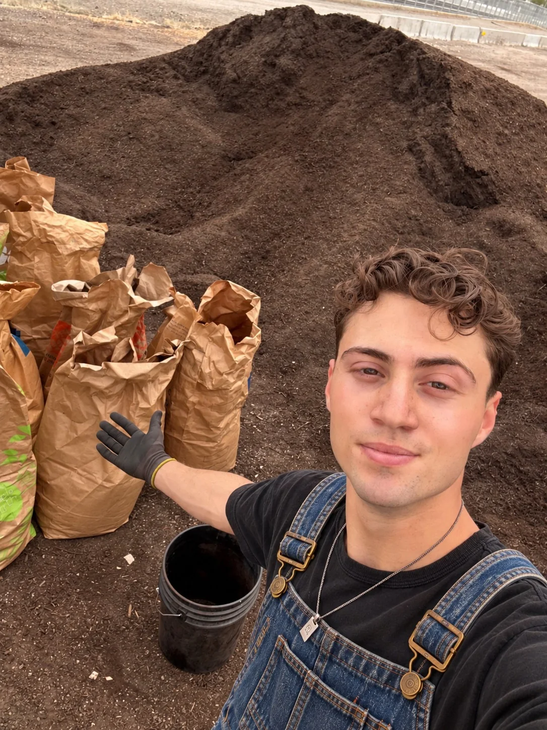 The real Ethan in overalls and work gloves, in front of a pile of finished compost and bags of food scraps